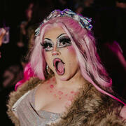 Portrait of Beth Amphetamine with her mouth open in a dramatic expression, exclaiming. She is wearing a large, glossy brown wig and looking past the camera.