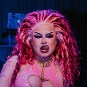 Portrait of Beth Amphetamine with her mouth open and tongue flicked out. Her eyes look to the side of the camera. She is wearing a large curly pink wig, swept back from her face.